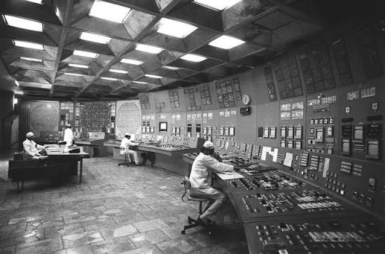 Black and white photograph of a Chernobyl control room: operators seated at a long console covered in hundreds of switches, with a wall of gauges and indicator panels behind them