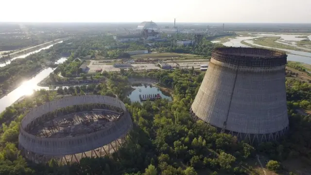 Aerial view of the Chernobyl site today: two unfinished cooling towers in the foreground amid dense forest, with the silver steel arch covering Reactor 4 in the background