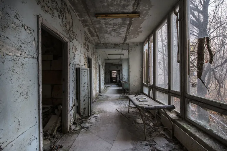 A corridor inside an abandoned building in Pripyat: paint peeling from the walls, debris across the floor, and broken windows looking out onto bare trees