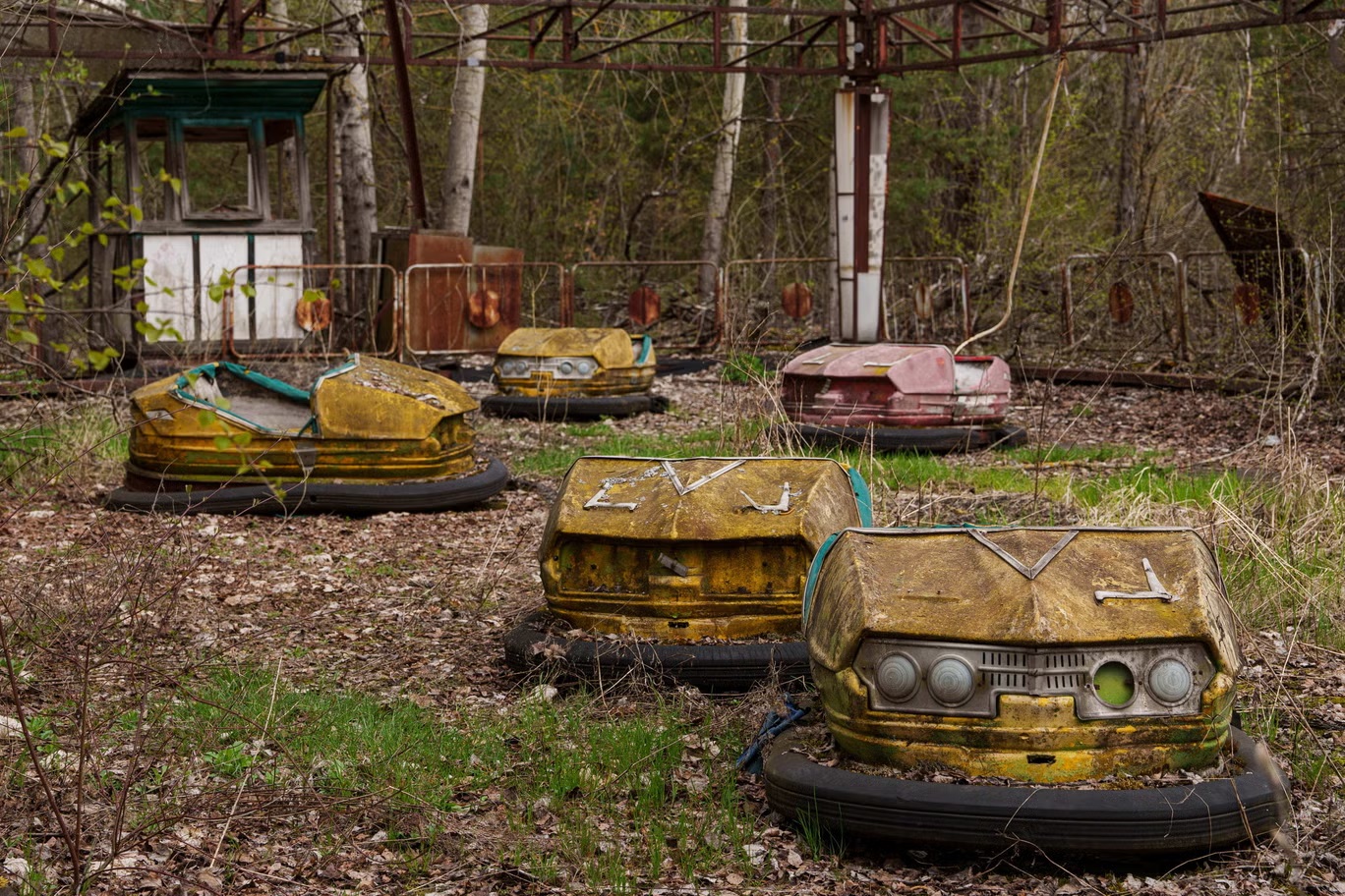 Rusted yellow bumper cars left in the Pripyat fairground, covered in moss and rust with grass growing around them among birch trees