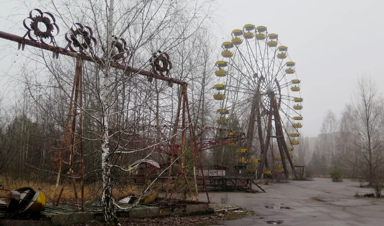 The yellow ferris wheel of abandoned Pripyat standing in fog, with a rusted swing carousel in front of it and trees growing up through the fairground