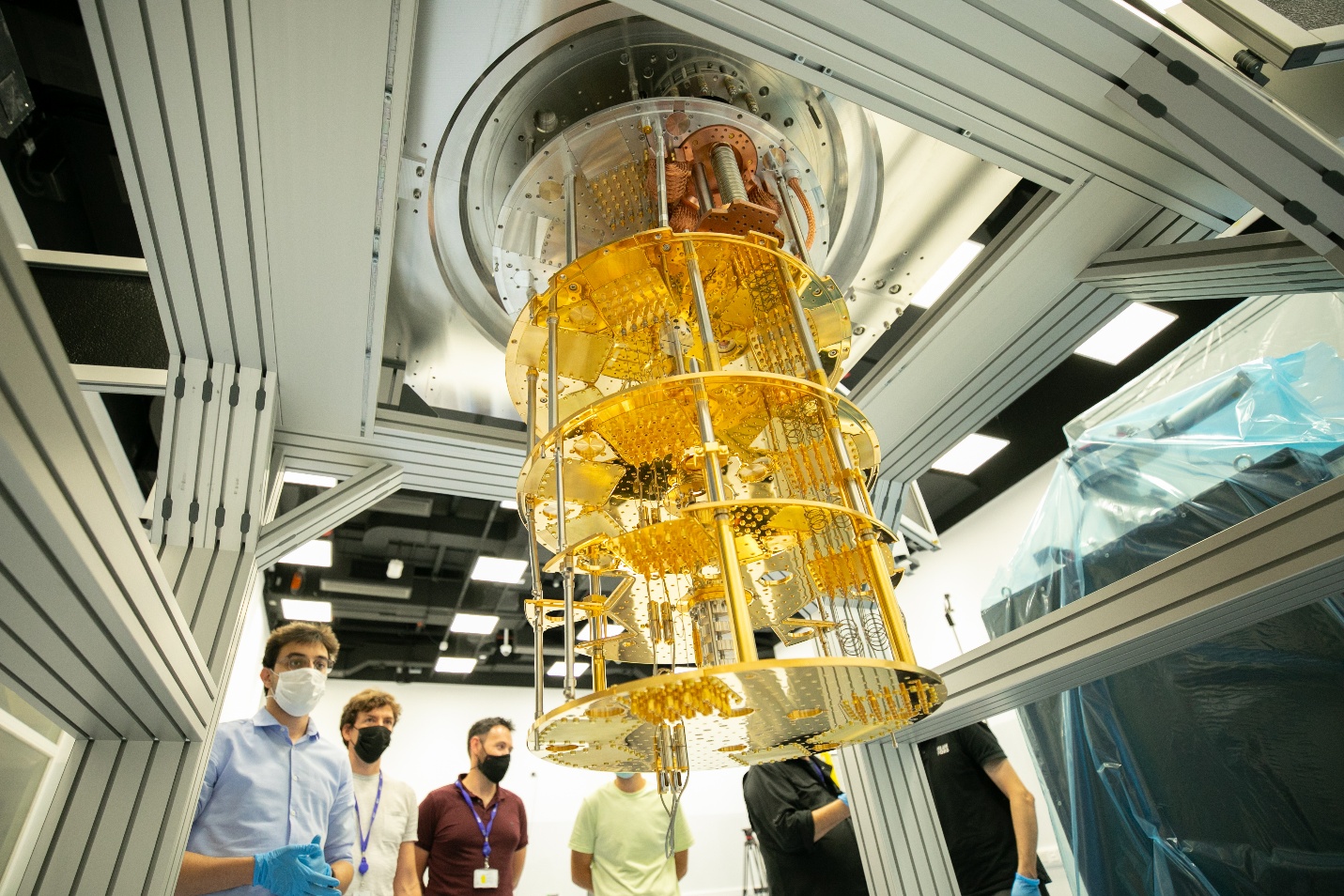 A quantum computer's dilution refrigerator suspended from the ceiling: stacked gold-coloured discs of decreasing size threaded with cables and pipes, with masked researchers standing beneath it
